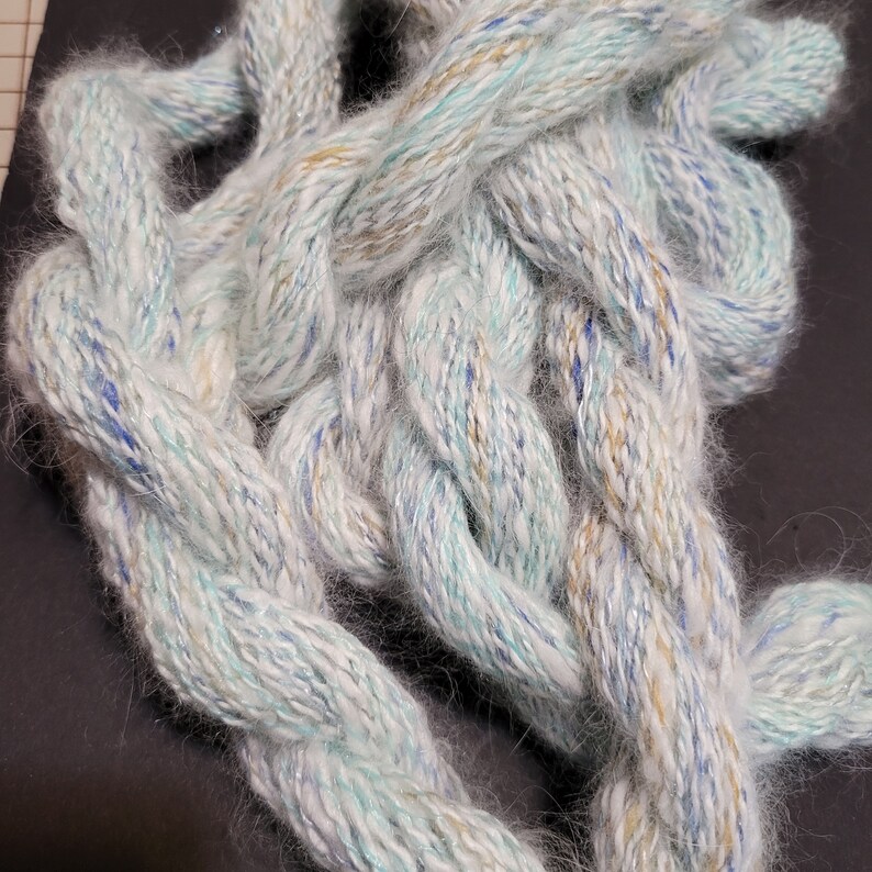 May include: A close-up of a white, blue, and gold yarn with a fuzzy texture. The yarn is twisted and knotted together.
