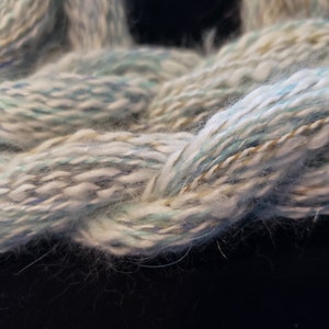 May include: Close-up of a soft, white yarn with blue, green, and gold accents. The yarn is fluffy and has a subtle shimmer.