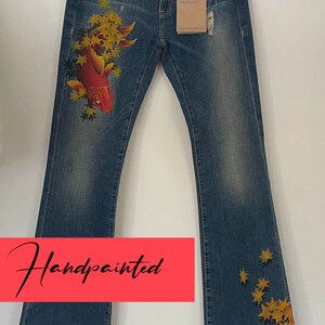 Puede incluir: Vaqueros de mezclilla azul pintados a mano con un diseño de pez rojo y hojas amarillas. Los vaqueros tienen un estilo de corte de bota y el texto "Handpainted" está escrito sobre un fondo rojo.