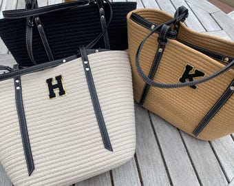 May include: Three rope-style tote bags in black, tan, and off-white. Each bag features black leather handles and straps with white stitching. The off-white and tan bags have a gold-trimmed initial on the front.