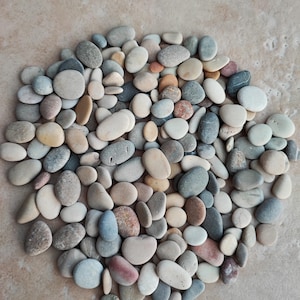 May include: A collection of smooth, oval river stones in various sizes and colours, including grey, beige, and reddish-brown. The stones are arranged in a circular pattern on a light brown surface, creating a natural, earthy aesthetic.