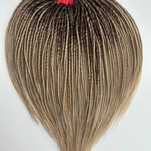 Puede incluir: Un conjunto de rastas sintéticas ombre marrón y rubio. Las rastas están atadas juntas con una cinta roja.