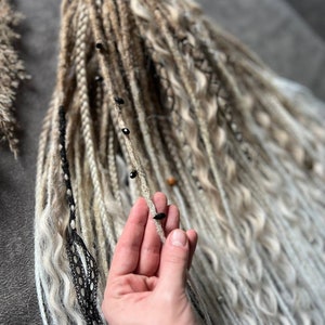 Puede incluir: Un conjunto de rastas sintéticas largas, blancas y marrones con cuentas negras. Las rastas se sostienen en una mano.