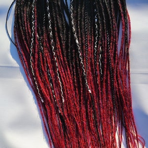 May include: A set of synthetic dreadlocks with a black to burgundy ombre colour. The dreadlocks are tied together with a brown string.
