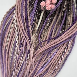 Puede incluir: Un paquete de rastas sintéticas en tonos de morado, rosa, marrón y blanco. Las rastas están atadas juntas con una cinta plateada y un pequeño ramo de flores rosas.