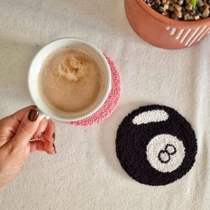 May include: A round punch needle coaster featuring a black and white 8-ball design, alongside a solid pink punch needle coaster supporting a white ceramic mug filled with a light brown beverage, and a terracotta planter with a green succulent.