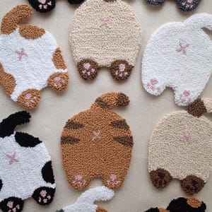 May include: A collection of punch needle coasters shaped like cat hindquarters in various colours including black, white, brown, tan, and calico patterns. Each coaster features a raised tail, two small feet with pink paw pads, and a pink stitched detail.