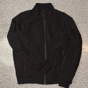 May include: Black bomber jacket with a zip-up front, a stand-up collar, and red accent stripes on the cuffs and hem. The jacket has a small pocket on the left sleeve and a red zipper pull on the chest pocket.