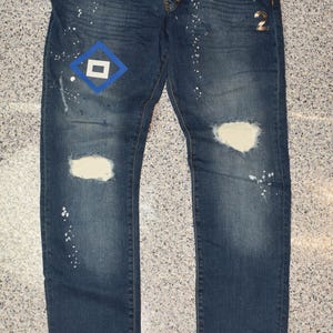 Pode incluir: Jeans azul escuro com detalhes desgastados, incluindo joelhos rasgados e respingos de tinta branca. Apresenta um patch em forma de diamante azul e branco e um detalhe dourado "2". Uma peça de roupa casual e elegante.
