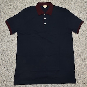 May include: Navy blue polo shirt with a burgundy collar and sleeve trim. The shirt has three white buttons and a small, gold-colored logo near the collar. The short-sleeved shirt is made of a soft material.