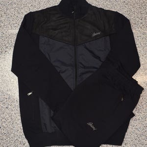 May include: A black track suit set with a jacket and matching pants. The jacket has a black suede-like panel across the shoulders and chest, with a zipper closure and the brand name "Brioni" embroidered on the chest. The pants have an elastic waistband.