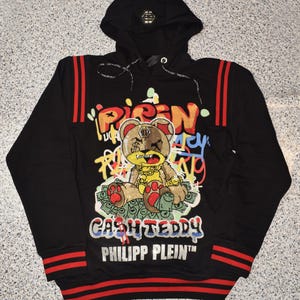 Puede incluir: Sudadera con capucha negra con rayas rojas y blancas en las mangas y la parte inferior. La sudadera presenta un oso de dibujos animados con una cadena de oro y sosteniendo dinero, con el texto "CASH TEDDY" y "PHILIPP PLEIN" debajo del oso.
