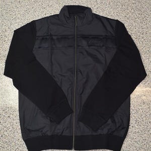 May include: A black zip-up jacket with a high collar. The jacket features black sleeves and a black body with a horizontal stripe design across the chest. The jacket has a small logo on the chest.