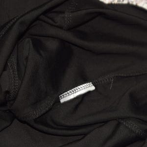 May include: Close-up of black fabric, possibly clothing, with visible stitching and a small white tag. The fabric appears soft and drapes loosely. The image focuses on the texture and construction of the garment.