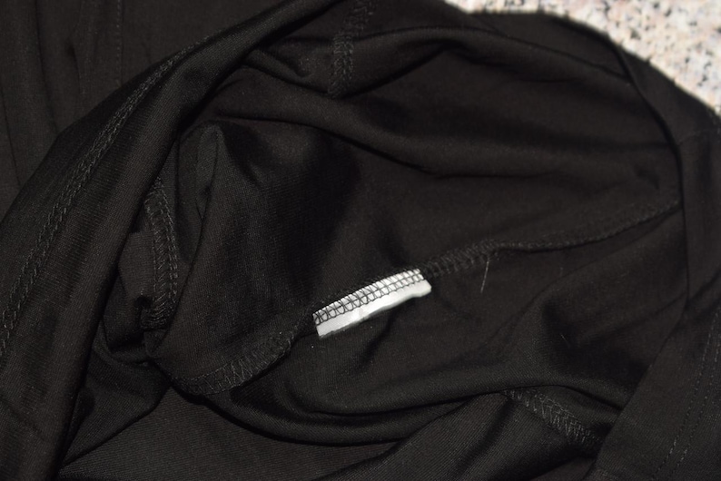 May include: Close-up of black fabric, possibly clothing, with visible stitching and a small white tag. The fabric appears soft and drapes loosely. The image focuses on the texture and construction of the garment.