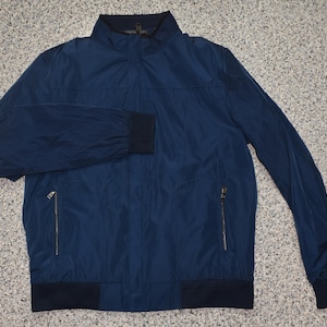 May include: A navy blue jacket with a stand-up collar and a zip closure. The jacket features two front zip pockets and ribbed cuffs and hem. The jacket is made of a lightweight material.