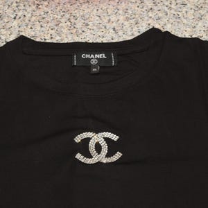 May include: Black t-shirt featuring a sparkling, interlocking double-C design. The shirt has a crew neck and a tag that reads "CHANEL". The XL size is visible on the tag. The shirt is made of a soft material.