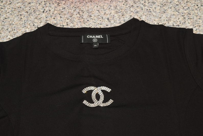 May include: Black t-shirt featuring a sparkling, interlocking double-C design. The shirt has a crew neck and a tag that reads "CHANEL". The XL size is visible on the tag. The shirt is made of a soft material.