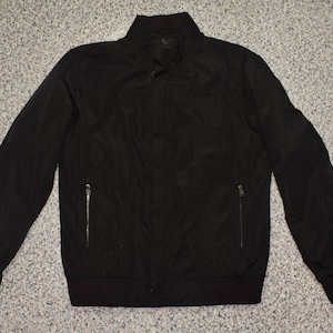 May include: A black jacket with a stand-up collar and two zippered pockets. The jacket has ribbed cuffs and a ribbed hem. The jacket is made of a smooth, lightweight material.