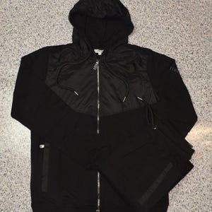 May include: Black hooded tracksuit set. The jacket has a quilted panel across the chest and a full zipper. The matching pants are folded neatly beside the jacket. The tracksuit is made of a soft material.