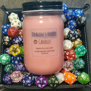 Może przedstawiać: R&oacute;żowa świeca w szklanym słoiku z czarną pokrywką. Świeca jest oznaczona jako "Dragon's Hoard Candles" i zawiera dwudziestościenną kostkę do gry. Słoik jest otoczony r&oacute;żnymi kolorowymi dwudziestościennymi kostkami do gry.