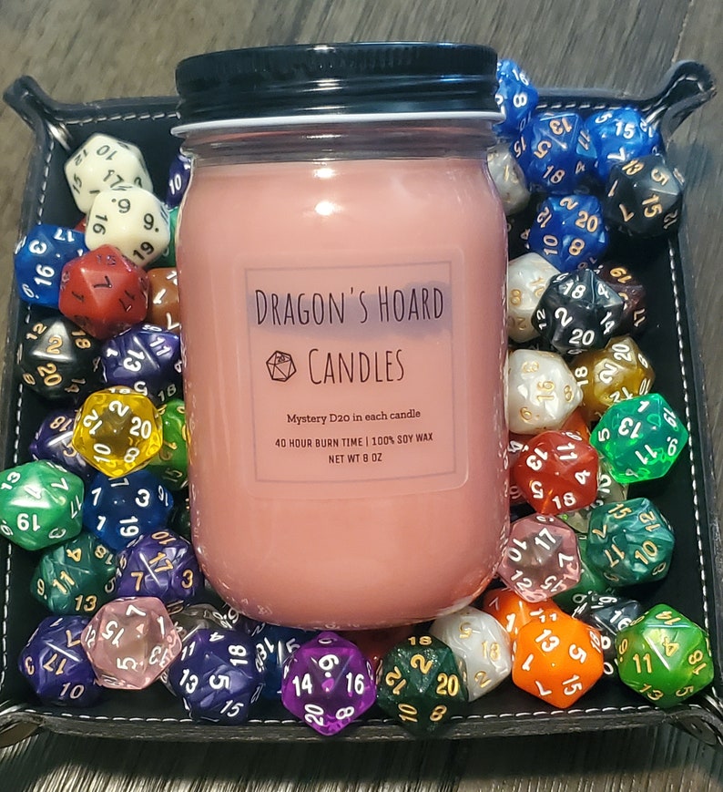 Może przedstawiać: R&oacute;żowa świeca w szklanym słoiku z czarną pokrywką. Świeca jest oznaczona jako "Dragon's Hoard Candles" i zawiera dwudziestościenną kostkę do gry. Słoik jest otoczony r&oacute;żnymi kolorowymi dwudziestościennymi kostkami do gry.