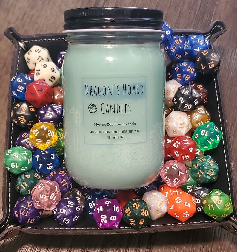 Może przedstawiać: Szklany słoik z tealową świecą, z czarną pokrywką i białą etykietą z napisem "Dragon's Hoard Candles". Etykieta zawiera r&oacute;wnież tekst "Mystery D20 in each candle", "40 Hour Burn Time | 100% Soy Wax" i "Net Wt 8 oz". Słoik ze świecą jest otoczony kolorowymi kostkami do gry.
