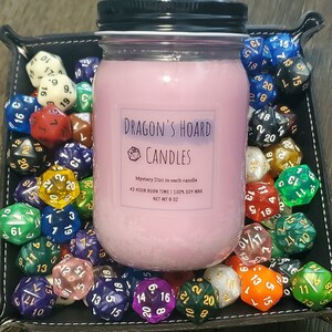 Może przedstawiać: R&oacute;żowa świeca sojowa w szklanym słoiku z czarną pokrywką. Słoik ma etykietę z napisem "Dragon's Hoard Candles" i zawiera tekst "Mystery D20 in each candle", "40 hour burn time | 100% soy wax" oraz "Net Wt 8 oz". Świeca jest otoczona kolorowymi dwudziestościennymi kostkami do gry.