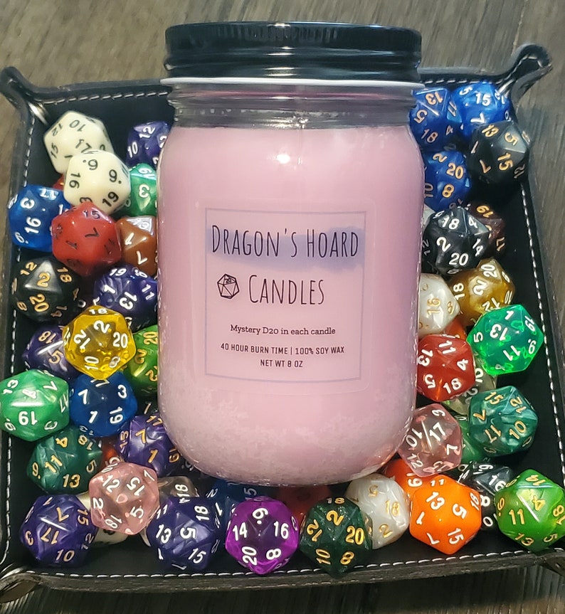 Może przedstawiać: R&oacute;żowa świeca sojowa w szklanym słoiku z czarną pokrywką. Słoik ma etykietę z napisem "Dragon's Hoard Candles" i zawiera tekst "Mystery D20 in each candle", "40 hour burn time | 100% soy wax" oraz "Net Wt 8 oz". Świeca jest otoczona kolorowymi dwudziestościennymi kostkami do gry.