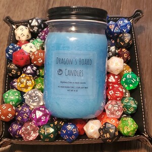 Może przedstawiać: Niebieska świeca sojowa w szklanym słoiku z czarną pokrywką. Słoik ma białą etykietę z napisem "Dragon's Hoard Candles" i zawiera informacje o świecy, takie jak czas palenia i rodzaj wosku. Świeca jest otoczona kolorowymi dwudziestościennymi kostkami do gry.