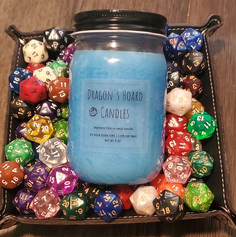 Może przedstawiać: Niebieska świeca sojowa w szklanym słoiku z czarną pokrywką. Słoik ma białą etykietę z napisem "Dragon's Hoard Candles" i zawiera informacje o świecy, takie jak czas palenia i rodzaj wosku. Świeca jest otoczona kolorowymi dwudziestościennymi kostkami do gry.