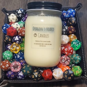 Może przedstawiać: Biały słoik ze świecą z czarną pokrywką i etykietą z napisem "Dragon's Hoard Candles". Etykieta zawiera r&oacute;wnież tekst "Mystery D20 in each candle", "40 hour burn time | 100% soy wax" i "Net Wt 8 oz". Słoik ze świecą jest otoczony r&oacute;żnymi kolorowymi kostkami do gry.
