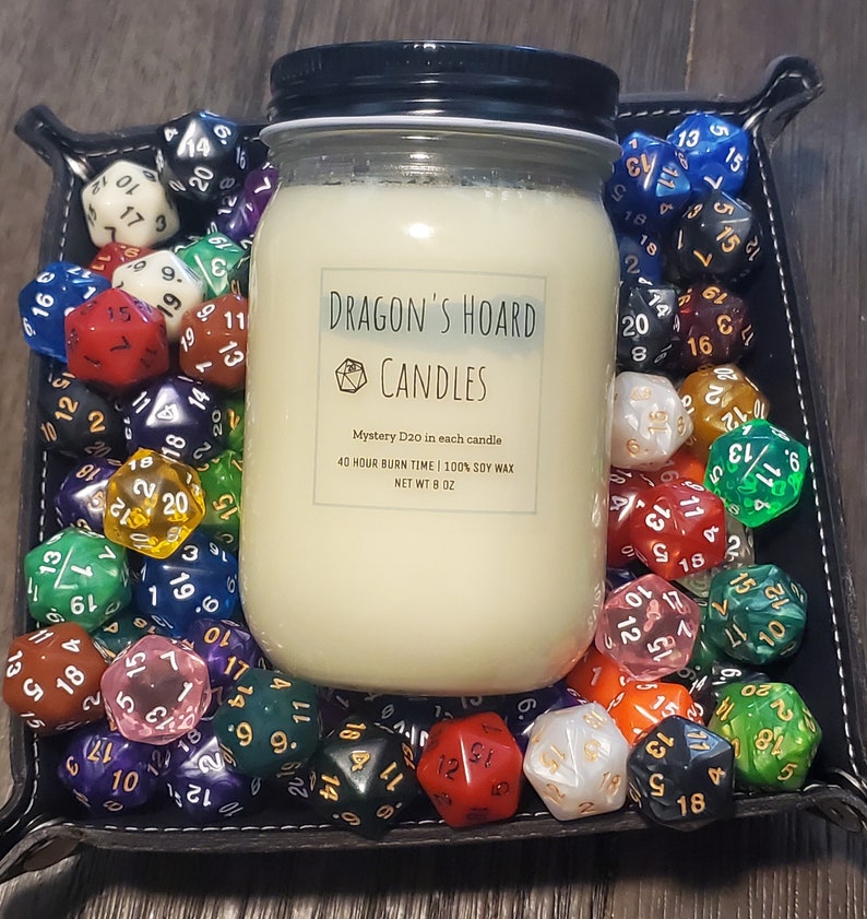 Może przedstawiać: Biały słoik ze świecą z czarną pokrywką i etykietą z napisem "Dragon's Hoard Candles". Etykieta zawiera r&oacute;wnież tekst "Mystery D20 in each candle", "40 hour burn time | 100% soy wax" i "Net Wt 8 oz". Słoik ze świecą jest otoczony r&oacute;żnymi kolorowymi kostkami do gry.