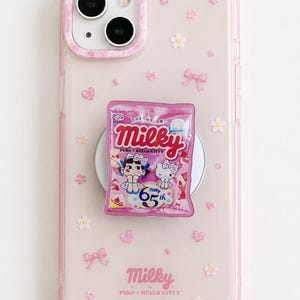 以下が含まれることがあります： ピンク色の透明な携帯電話ケース。ピンクと白の花柄とリボンのデザイン。 「Milky」と「Peko x Hello Kitty」の文字が入ったピンク色のキャンディーパッケージが携帯電話ケースの裏側に取り付けられています。