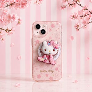 以下が含まれることがあります： ピンクの桜模様とHello Kittyの文字がデザインされた透明なスマートフォンケース。ピンクの柄入り着物を着て、花柄の髪飾りをつけたHello Kittyの立体的なフィギュアがケースの中央に取り付けられています。