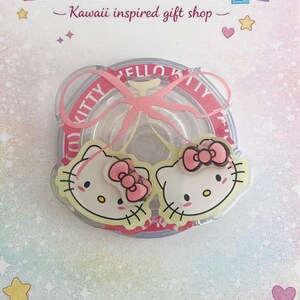 Può includere: Un accessorio circolare trasparente con due ciondoli a forma di viso di Hello Kitty con fiocchi rosa. L'accessorio è circondato da un nastro rosa e dalla scritta "HELLO KITTY". La parte superiore dell'immagine recita "Kittycutiedesign - Kawaii inspired gift shop".