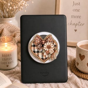 Puede incluir: Un lector electrónico Kindle negro con un accesorio de agarre circular en la parte posterior. El accesorio muestra un diseño en forma de corazón con un patrón de cuadros negro y beige, un lazo de leopardo y flores de tela en tonos crema.