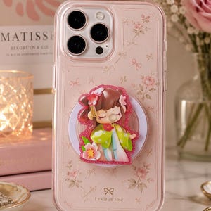 Op de afbeelding: Een transparant telefoonhoesje met een lichtroze bloemenpatroon en een klein strikicoontje boven de tekst La vie en rose. In het midden is een ronde telefoonhouder bevestigd met een stripfiguur met bruin haar in een groen-wit outfit.