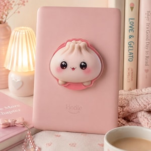 Puede incluir: Un lector electrónico Kindle de color rosa claro con un agarre tridimensional centrado en forma de dumpling. El agarre muestra una cara de dibujos animados con ojos negros grandes, mejillas sonrosadas y una boca pequeña. La palabra kindle es gris.