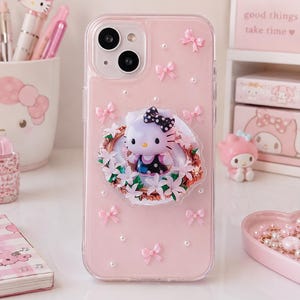 Op de afbeelding: Een doorzichtig telefoonhoesje met een lichtroze achtergrond, versierd met kleine witte parels en roze strikjes. In het midden bevindt zich een driedimensionaal Hello Kitty-figuurtje, omlijst door een goudkleurige rand met lichtroze bloemen.