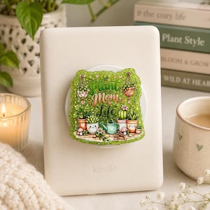 Op de afbeelding: Een witte e-reader met een glinsterende, rechthoekige zelfklevende grip op de achterkant. De grip toont een groene illustratie van diverse potplanten, een gieter en de tekst "Plant Mom Life" in een decoratief schrijflettertype.