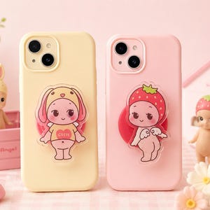 Puede incluir: Dos fundas para teléfono, una amarillo pálido y otra rosa, cada una con un diseño de personaje de dibujos animados. La funda amarilla presenta un personaje de conejito, y la funda rosa tiene un personaje de fresa. Ambas fundas tienen un acabado brillante.