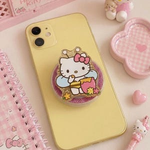 以下が含まれることがあります： ハローキティをテーマにしたスマホグリップが付いた黄色のスマートフォン。ピンクのリボン、青い羽、蜂蜜の壺を持つ漫画のキャラクターが特徴です。その他のピンク色のアクセサリーには、メモ帳、ペン、置物などがあります。