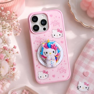 以下が含まれることがあります： ピンク色のスマートフォンケース。ハローキティのデザインで、虹色のたてがみとピンクのリボンが付いた3Dハローキティのフィギュアが特徴です。ケースには「Hello Kitty」の文字やその他のハローキティのグラフィックも描かれています。