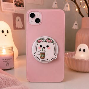 Op de afbeelding: Een lichtroze smartphonehoesje met een ronde telefoonhouder. De houder toont een cartoonspookje met een roze bloemenkrans dat een ijskoffie met een groen rietje vasthoudt. Het hoesje heeft een uitsparing voor de dubbele camera linksboven.