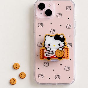 以下が含まれることがあります： ハローキティのデザインが施されたピンク色の携帯電話ケース。キャラクターがクッキーを持っています。ケースは透明な縁取りで、ハローキティの顔と小さな黒いハートで飾られています。3つの小さくて丸いクッキーが近くに散らばっています。