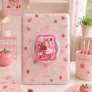 May include: A light pink Kindle e-reader case featuring a repeating pattern of small red strawberries, white flowers, and pink bows. A rectangular pop-out grip is attached to the centre, displaying an illustration of a white goose with a basket.