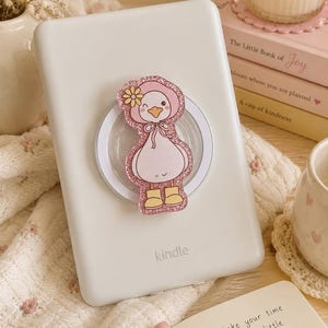 Può includere: Un lettore e-reader Kindle bianco con un supporto circolare per telefono sul retro. L'accessorio presenta un'illustrazione glitterata in stile cartone animato di un'anatra bianca con una cuffietta rosa, un fiore giallo e stivali gialli.