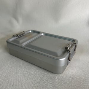 Puede incluir: Una fiambrera rectangular de metal plateado con tapa con bisagras y cierre seguro. El interior está dividido en dos compartimentos. La fiambrera está diseñada para transportar alimentos y es adecuada tanto para adultos como para niños.