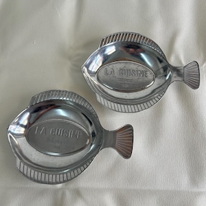 May include: Two silver-colored, fish-shaped serving dishes. Each dish has a textured rim and a tail fin. The oval center of each dish is imprinted with the words "LA CUISINE DEPUIS 1958".